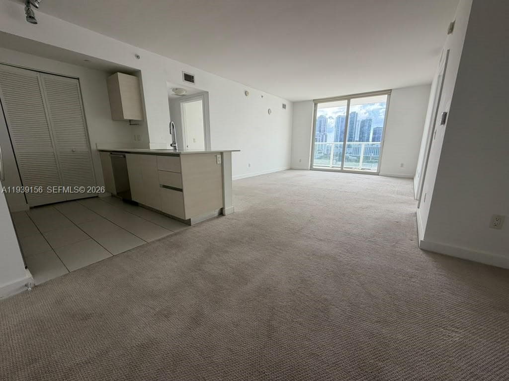 Open-concept interior featuring light-toned cabinetry, a peninsula countertop, ceramic tile flooring in the kitchen, wall-to-wall carpeting, and floor-to-ceiling glass sliding doors