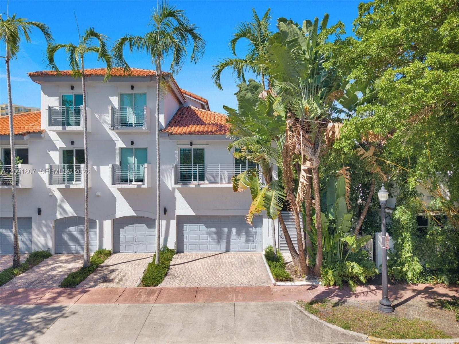 Three-story exterior featuring a terracotta tile roof, white stucco facade, multiple private balconies with metal railings, and attached multi-car garages with paver driveways
