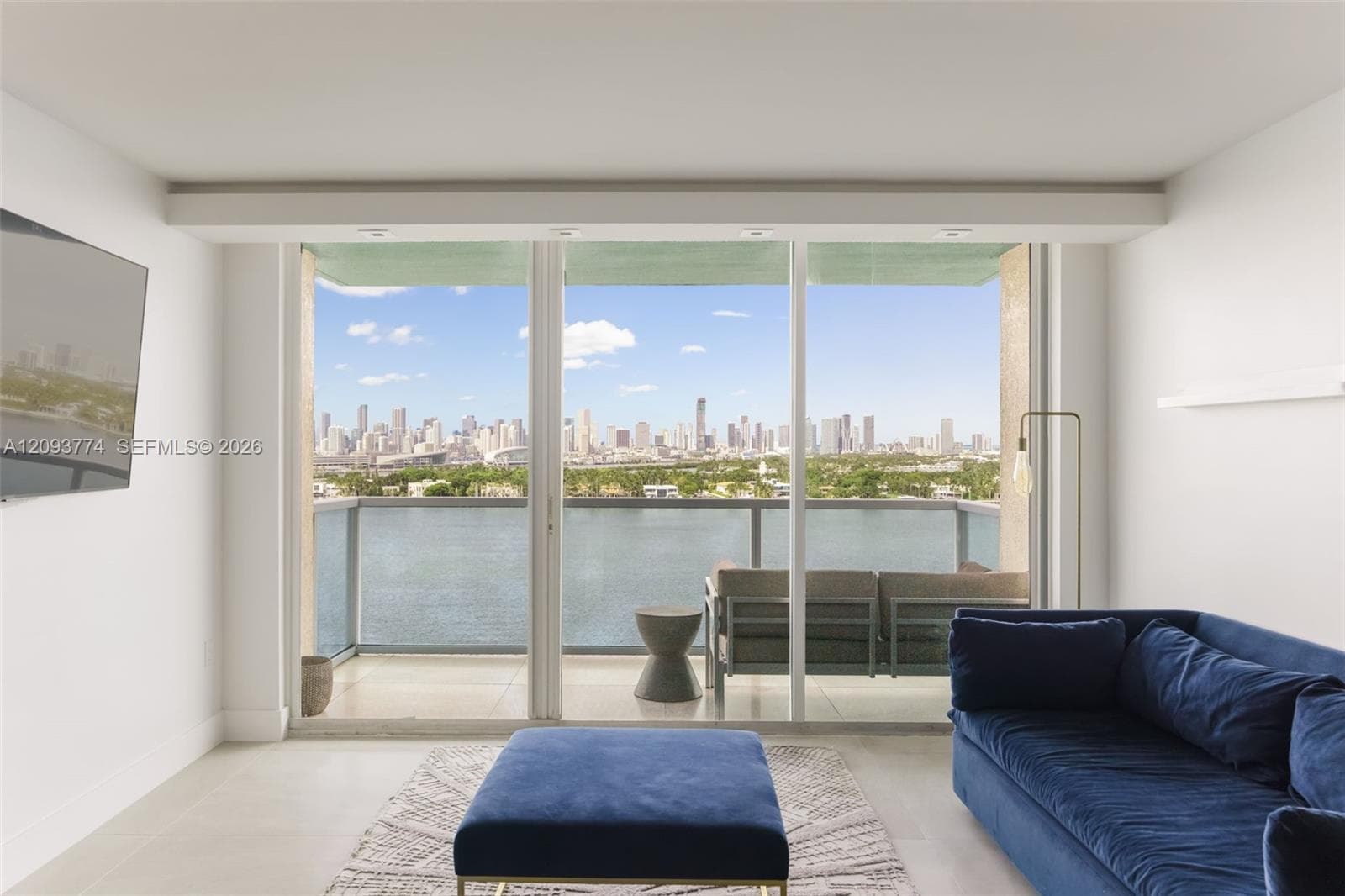 Floor-to-ceiling glass doors, private balcony with glass railing, large-format tile flooring, and panoramic cityscape and waterfront backdrop