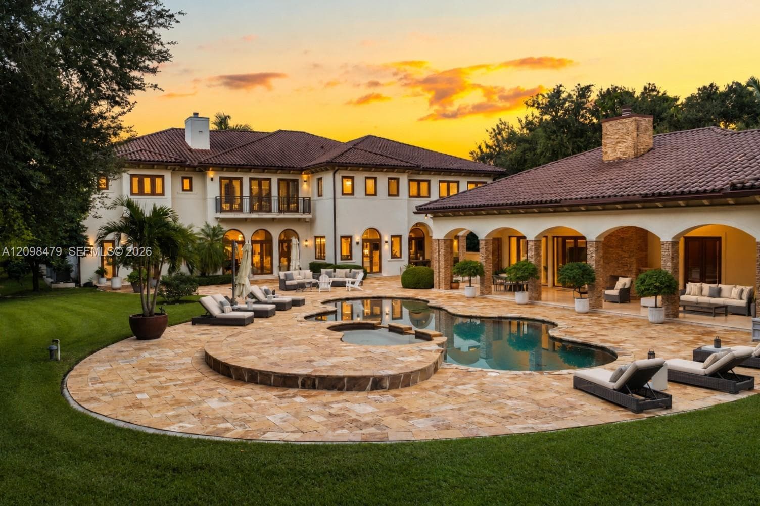 Stucco exterior with multi-level tile roofing, expansive stone-paved pool deck, freeform swimming pool with raised spillover spa, and covered loggia featuring stone-clad pillars and a built-in outdoor fireplace