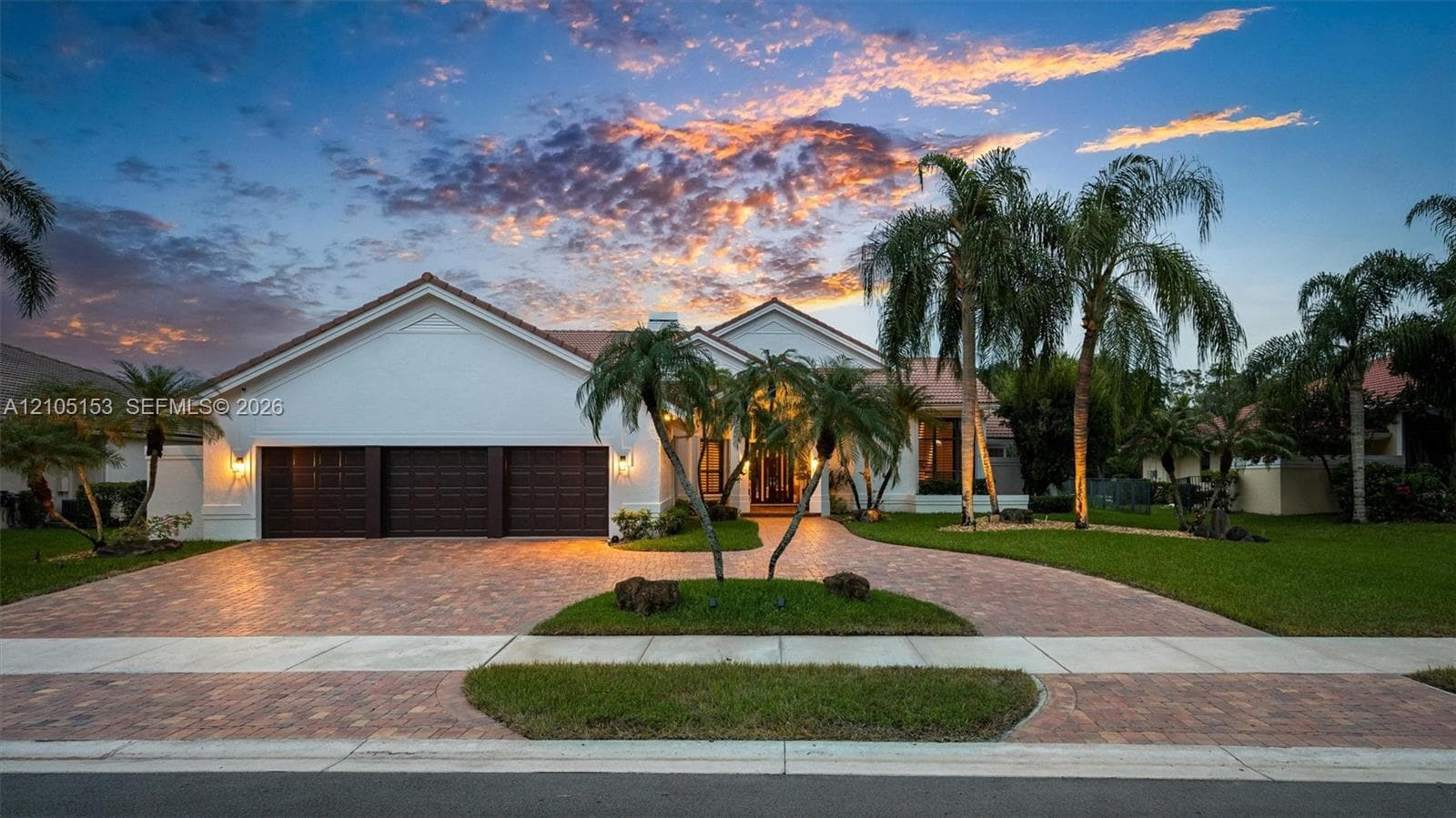 Stucco exterior with a tile roof, paver driveway, three-car garage, and established palm trees