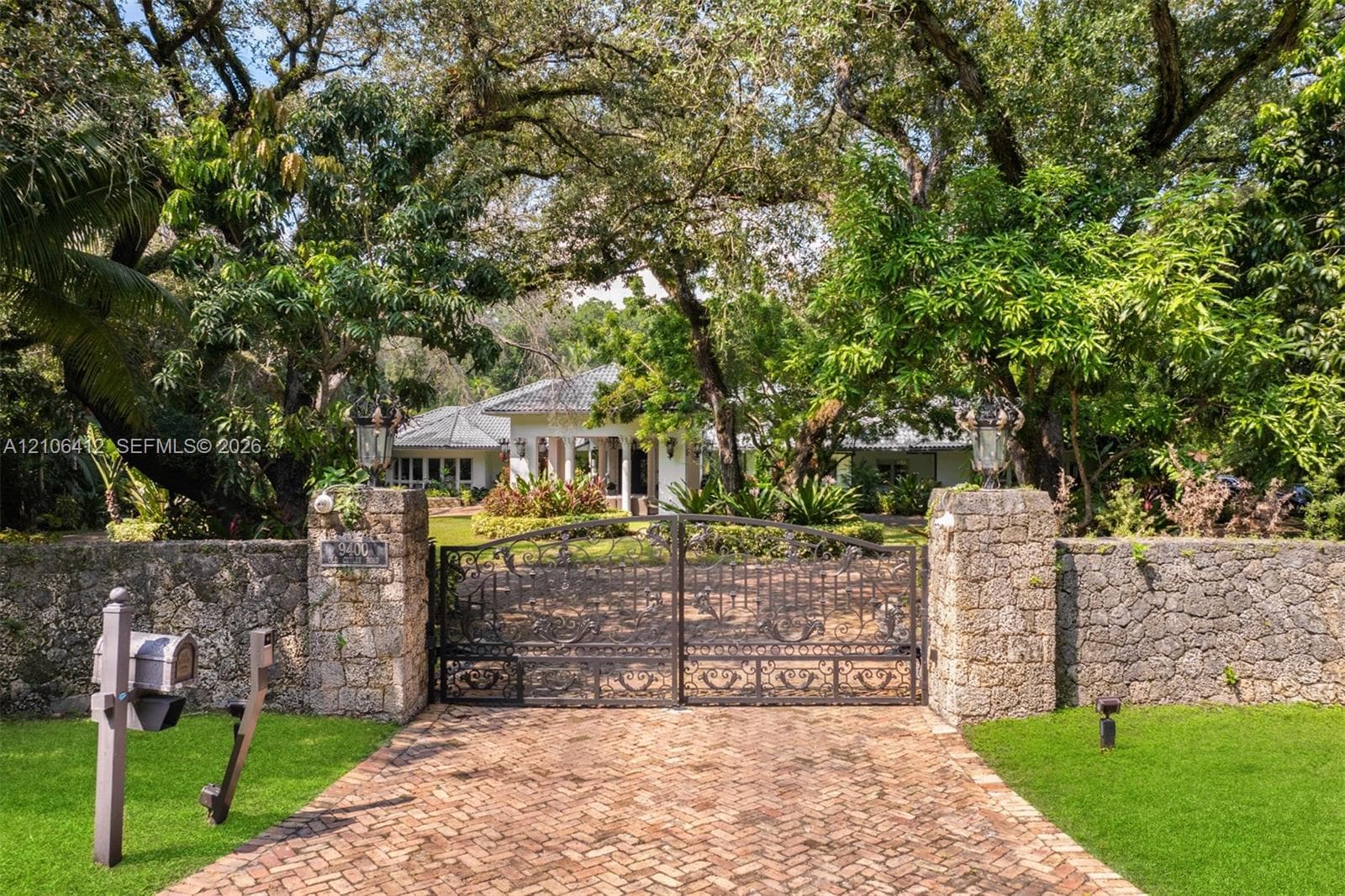 Gated estate entrance featuring a herringbone-patterned brick driveway, stone perimeter wall, ornamental metal gate, and mature canopy trees