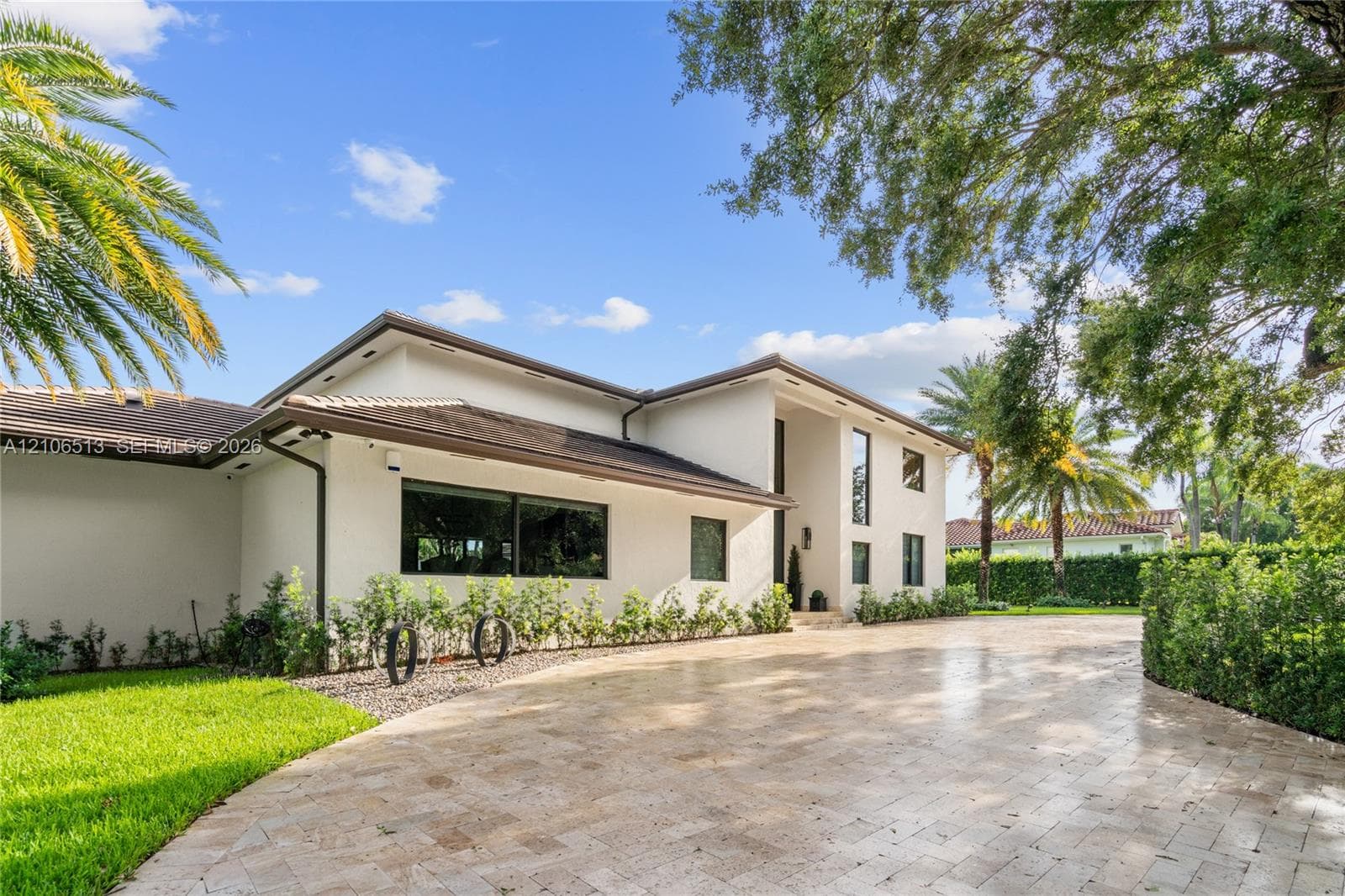 Stucco exterior featuring a low-pitch roof, expansive stone-paver driveway, and mature landscaping