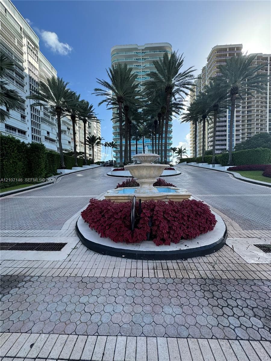 Gated entrance featuring a tiered stone fountain, stone paver driveway, mature palm trees, and manicured landscaping