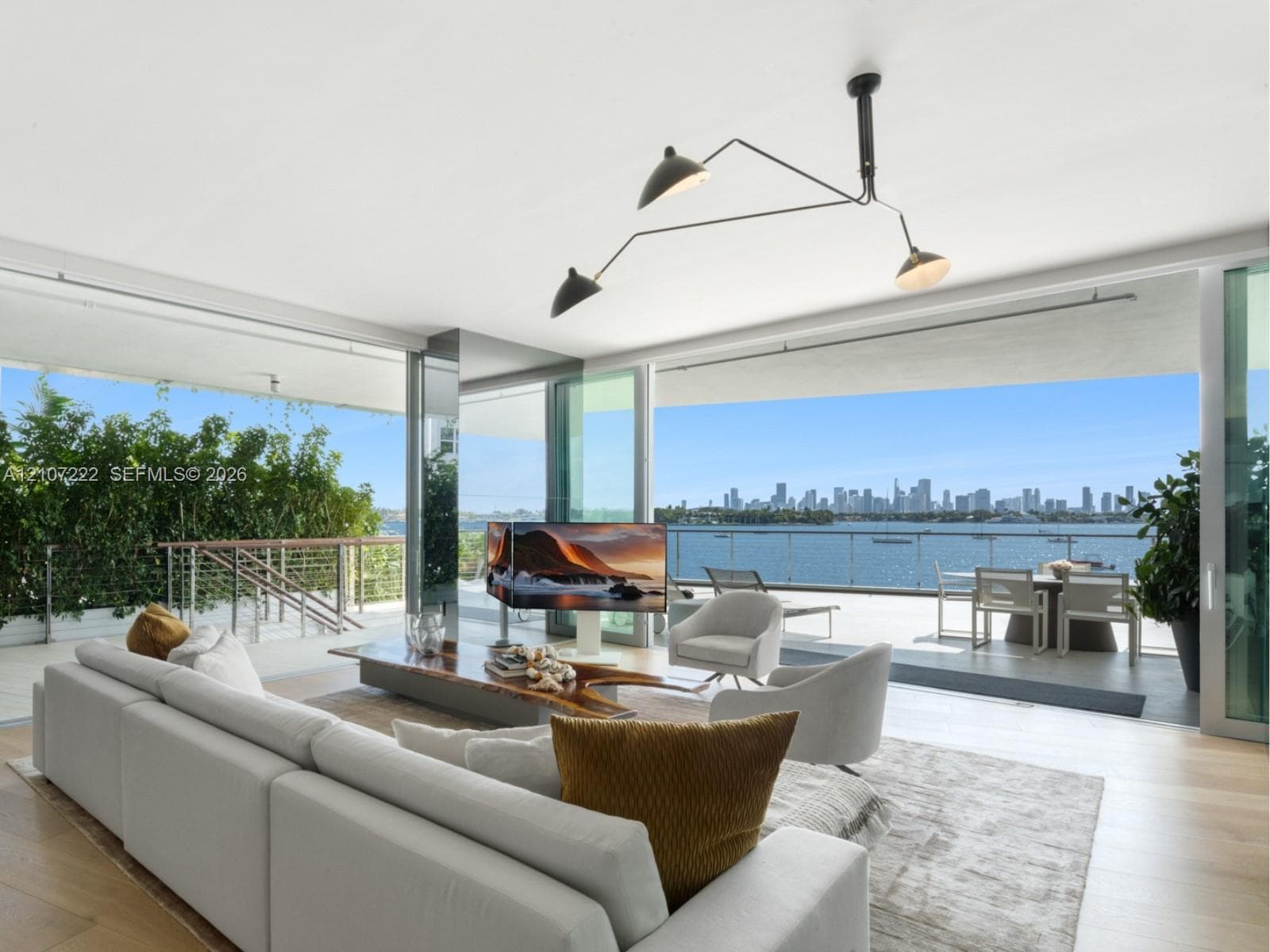 Open-concept interior featuring floor-to-ceiling glass sliding doors, wood-finish flooring, architectural multi-arm pendant lighting, and a wrap-around balcony with metal railing