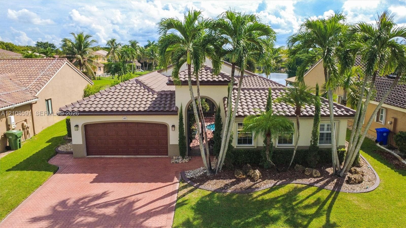 Stucco exterior featuring a clay tile roof, arched entryway with ornamental iron gate, two-car garage with paneled door, paver driveway, and mature palm trees