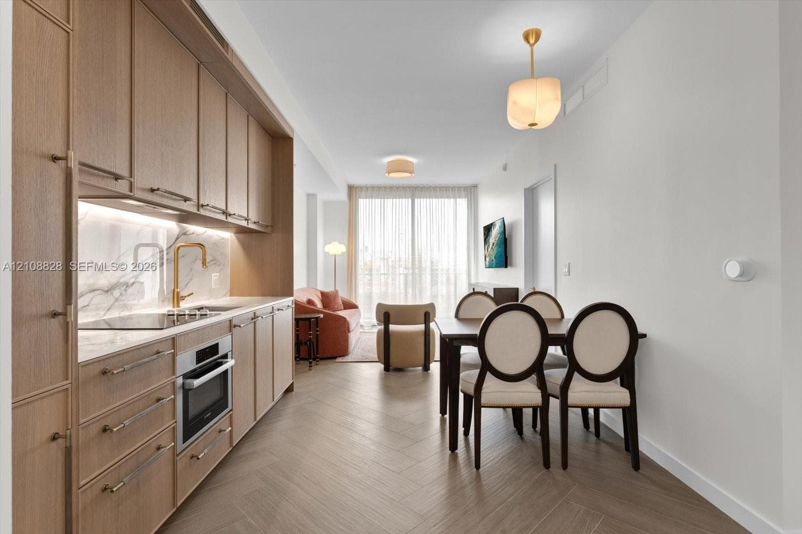 Kitchen featuring light wood-finish cabinetry, integrated appliances, stone-surface backsplash, and gold-toned hardware, complemented by herringbone-pattern flooring